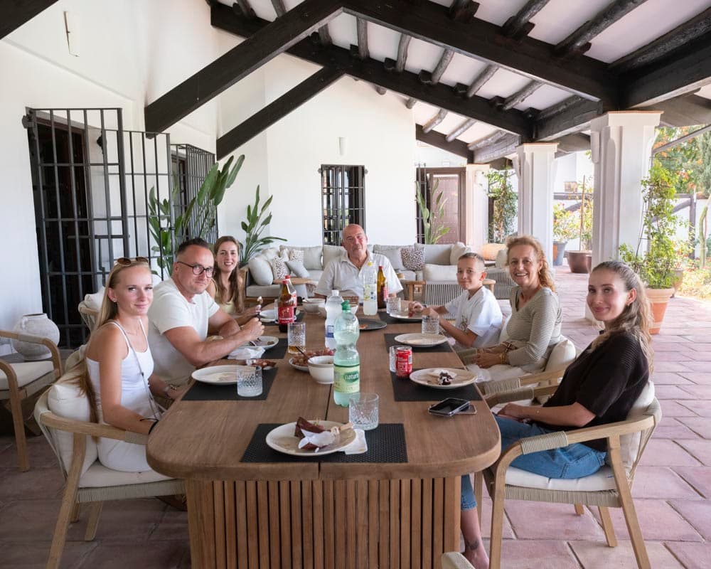 Grupo familiar almorzando en el porche cubierto de la casa, con vigas de madera oscura, mesa de teca natural y sillas de ratán con cojines blancos. Al fondo se aprecia la zona de sofás exteriores y el jardín.
