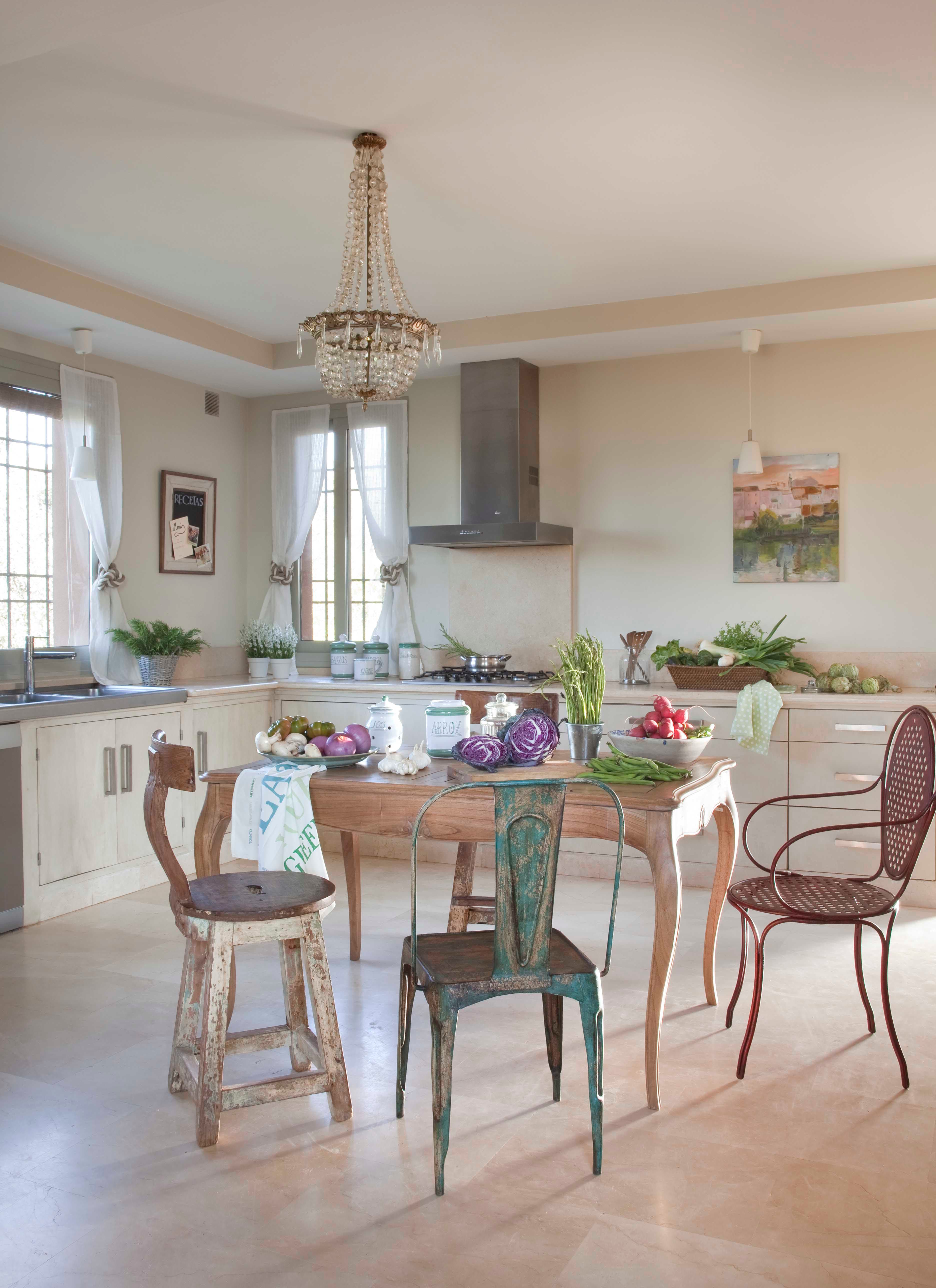 Cocina de estilo campestre con muebles en blanco roto, isla central de madera con cesta de mimbre para botellas y encimeras de piedra gris. Una araña de cristal preside el centro. Las sillas son un ecléctico mix de estilos —de hierro vintage, madera envejecida y rejilla— agrupadas alrededor de la isla cargada de verduras frescas.